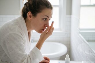 Jeune femme posant une lentille de couleur sur son doigt devant un miroir de salle de bain, avec étui et solution lentilles visibles